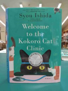 Photo of the cover for the novel 'Welcome to the Kokoro Cat Clinic', featuring a black cat wearing a doctor's head mirror and two miniature cats playing in pill bottles