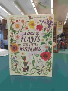 Photo of the cover for the junior nonfiction book 'A Guide to Plants for Little Witchlings', featuring a variety of illustrated flowers, with a small child and cat looking up at them